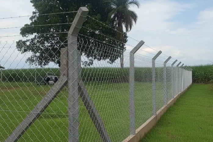 Alambrado galvanizado de malha losango sobre base de concreto, com braços inclinados de arame farpado, cercando área rural com canavial ao fundo