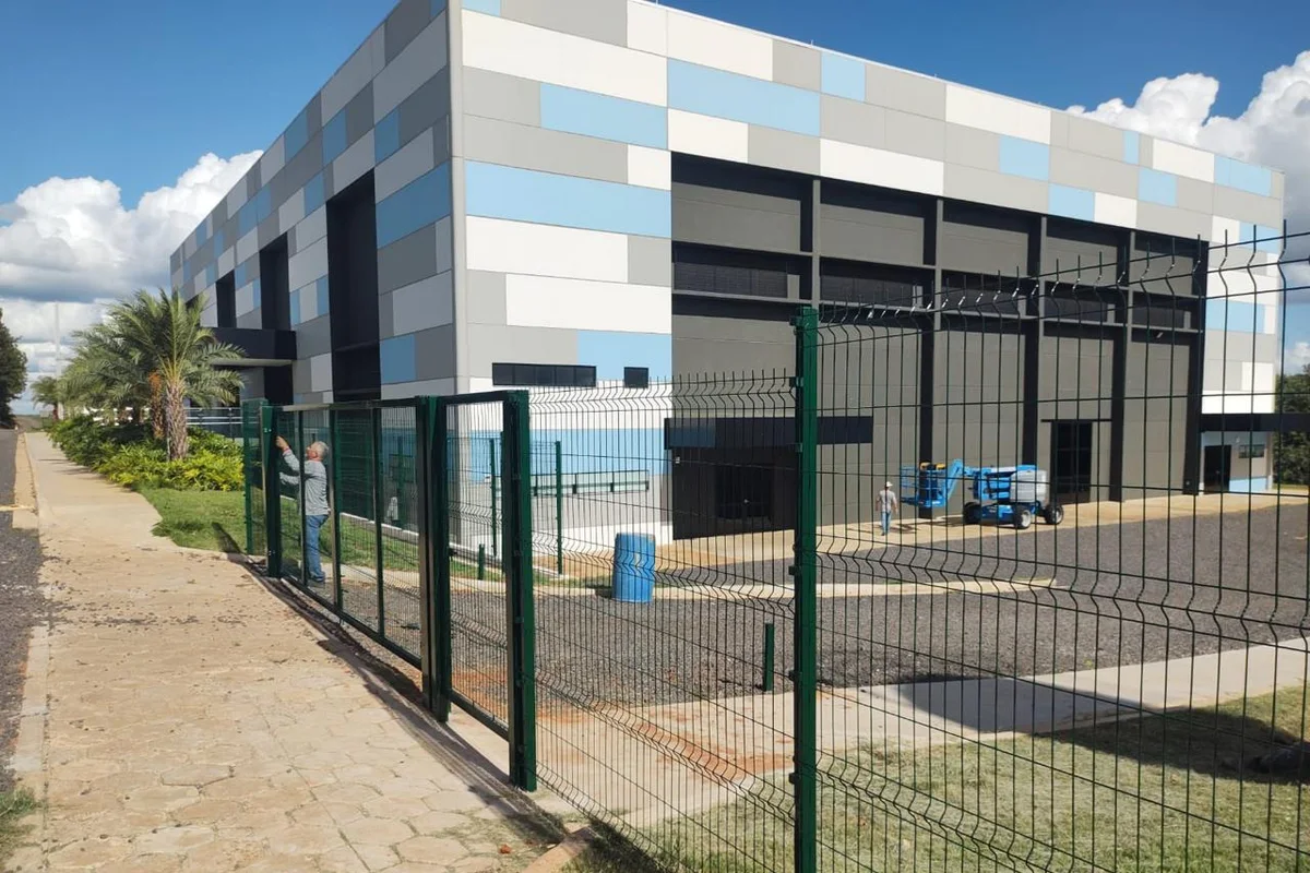 Gradil de painel soldado pintado de escuro instalado na fachada de um galpão industrial, com portão de acesso e passeio de concreto