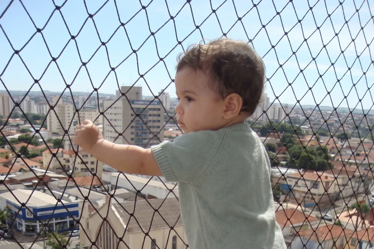 Criança em pé na sacada de um apartamento, protegida por rede de proteção instalada de ponta a ponta do guarda-corpo